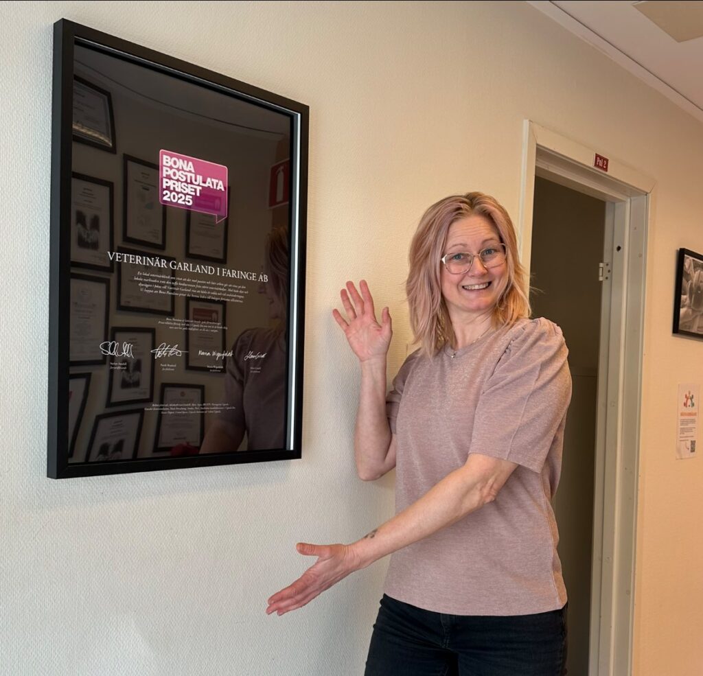 Jenny, iklädd en svagt rosa tunika och svarta byxor, står intill en vit vägg på kliniken och håller upp armarna och pekar mot en tavla på väggen. Tavlan med svart ram innehåller motiveringen till att Jenny fick priset Bona Postulata.